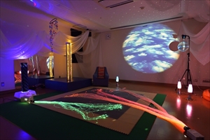 スヌーズレン用の部屋の画像。スクリーンに空が映し出され、様々な色のライトや布等で装飾がされている。